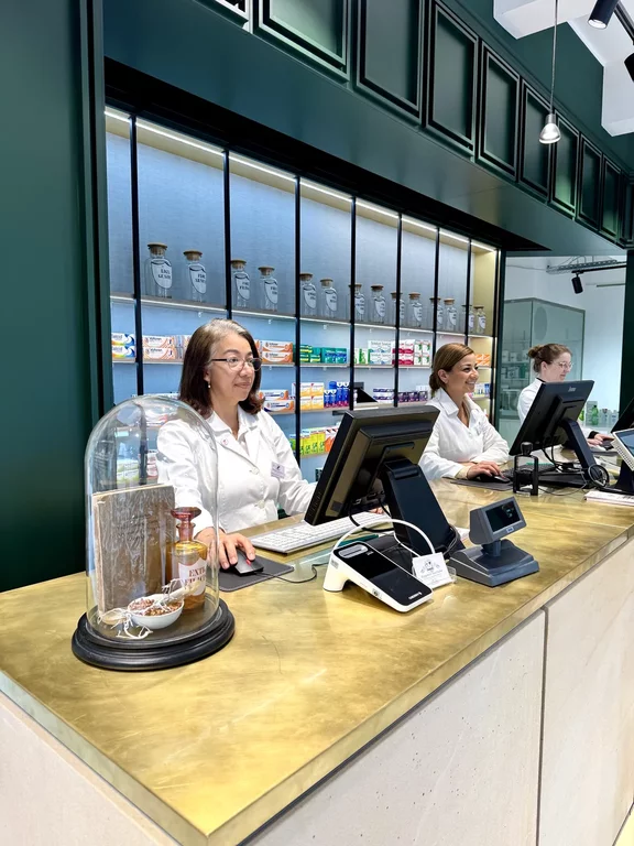Offizin der Elefanten-Apotheke im Jubiläumsjahr 2025. Die Einrichtung ist tannengrün, die Oberfläche des Verkaufstisches messing-gold, dahinter stehen 3 Mitarbeiterinnen.